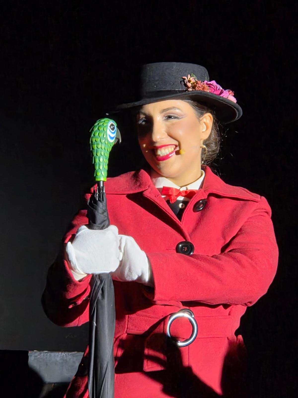Attrice in scena nei panni di Mary Poppins, con cappello, cappotto rosso e ombrello